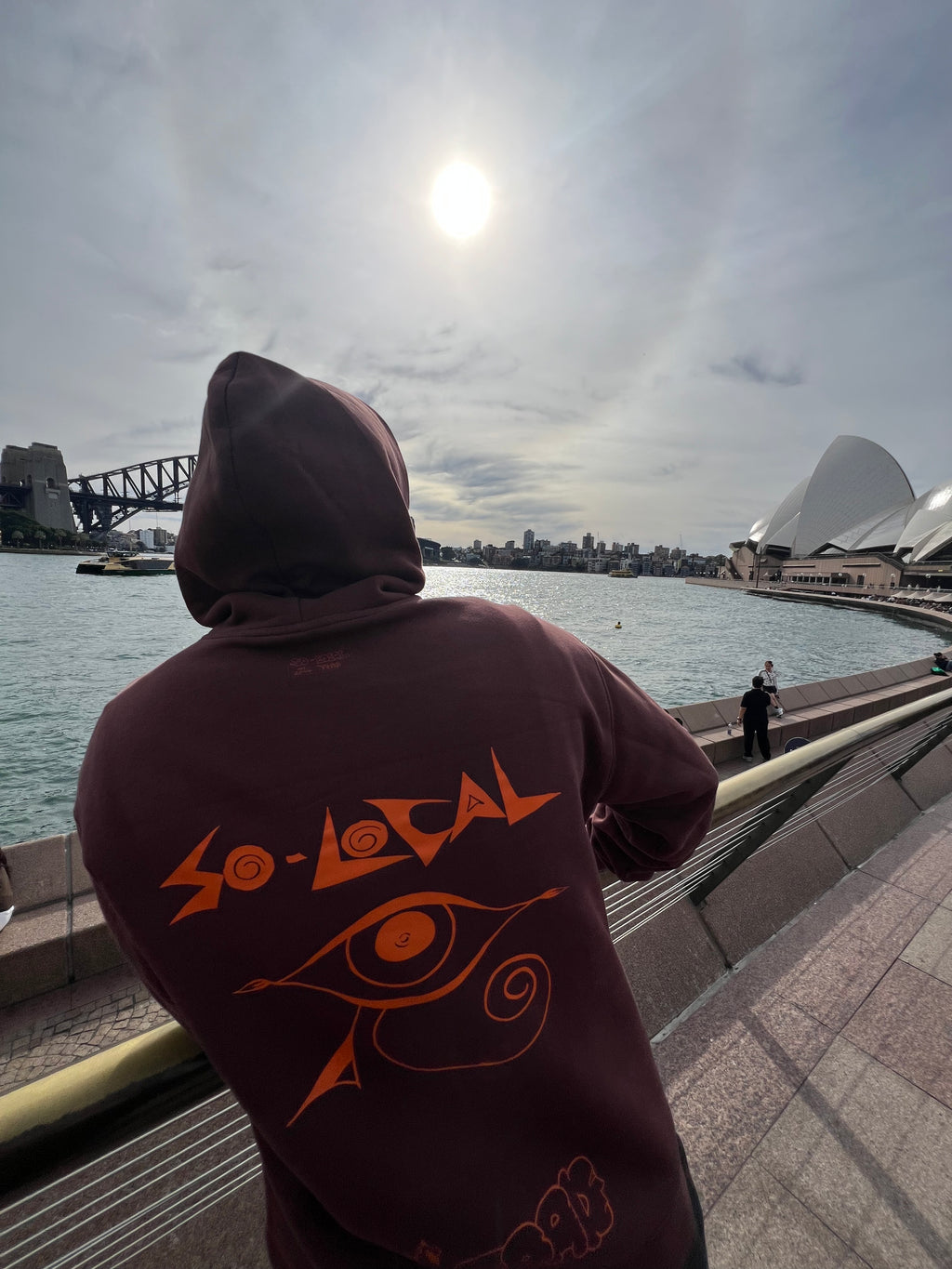 "I SEE YOU" SO LOCAL brown hoodie back print, worn overlooking Sydney Opera House and harbour