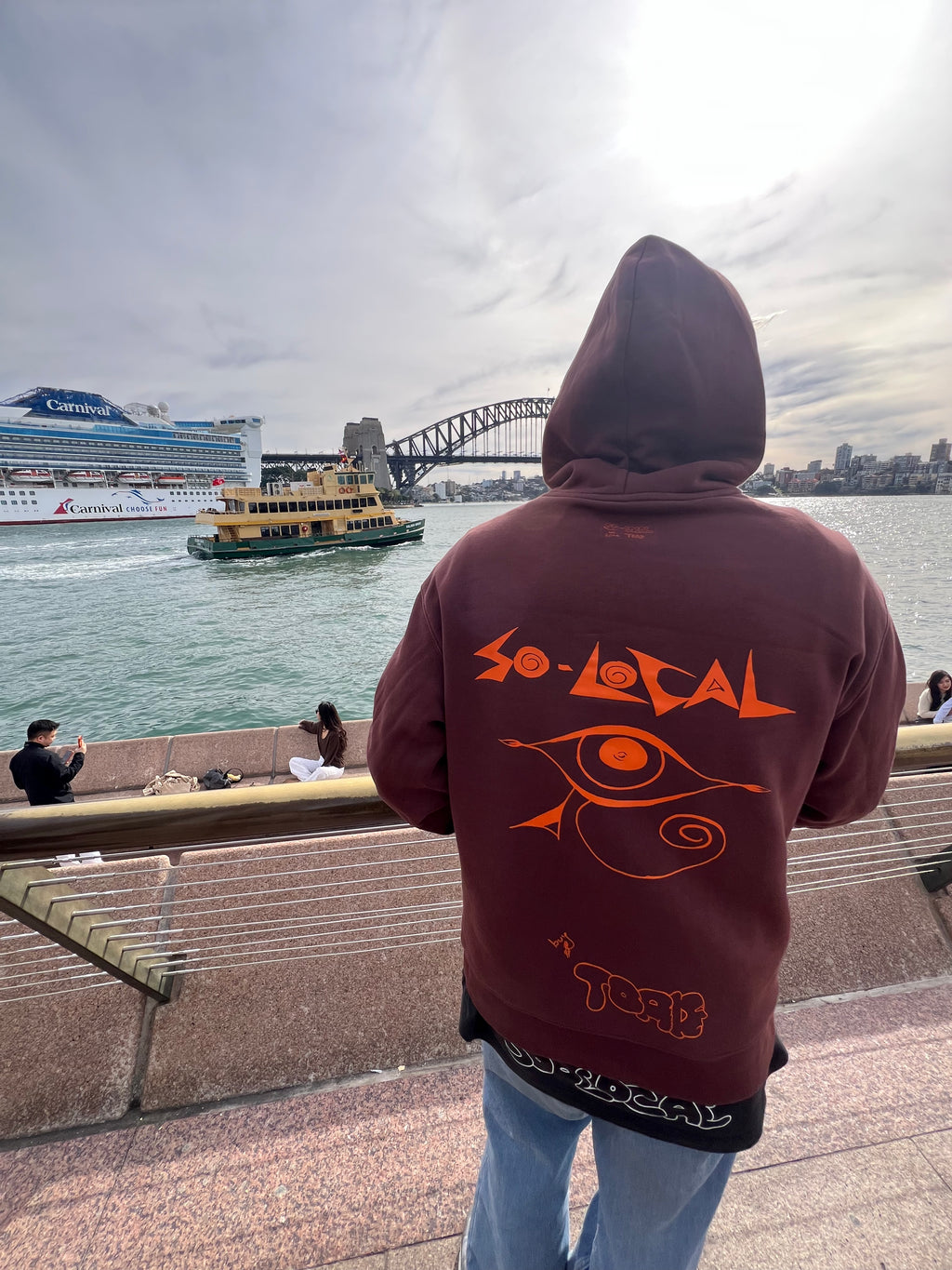"I SEE YOU" SO LOCAL brown hoodie back print, worn at Sydney Harbour with cruise ship and ferry in background