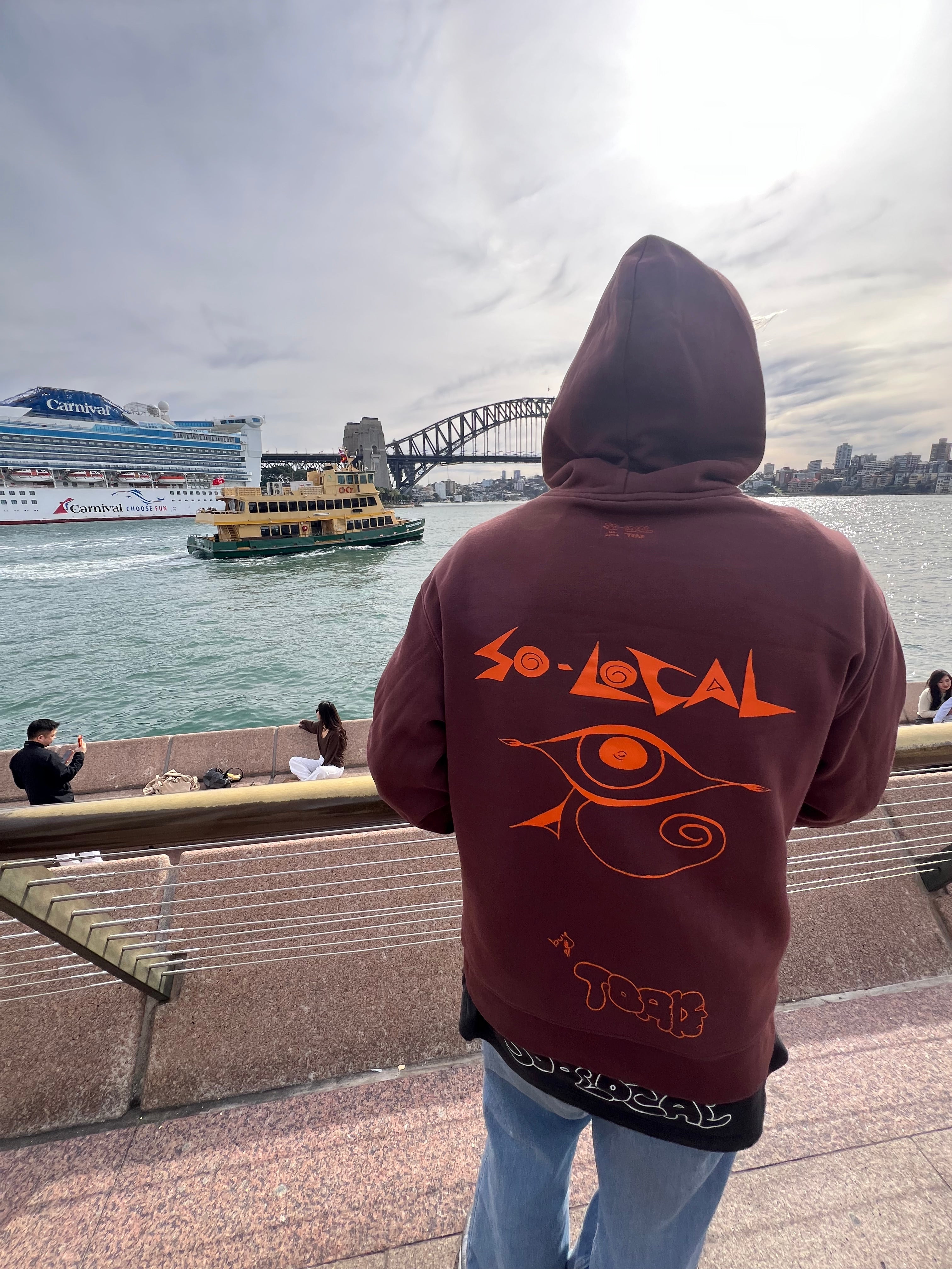 "I SEE YOU" SO LOCAL brown hoodie back print, worn at Sydney Harbour with cruise ship and ferry in background