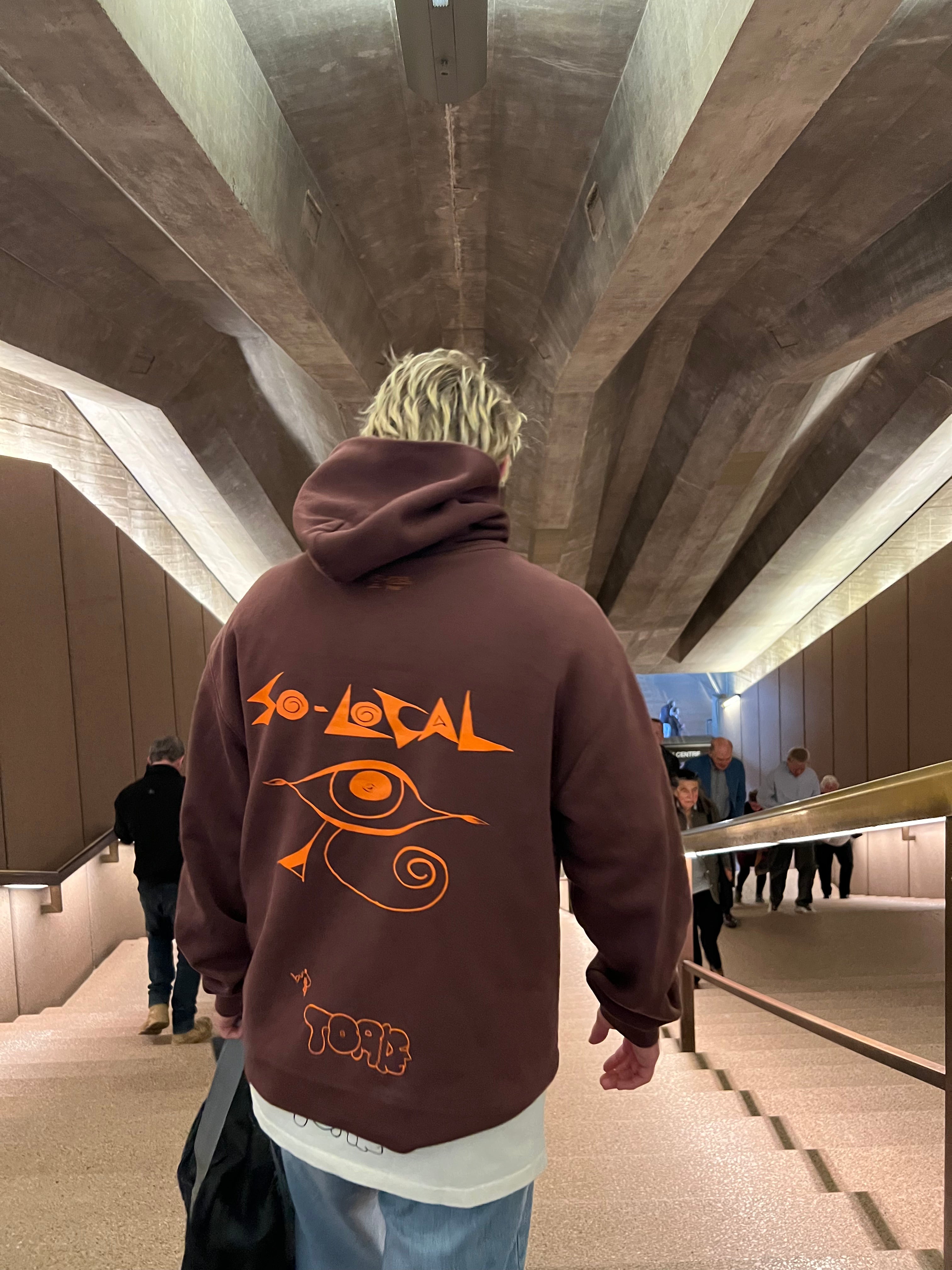 "I SEE YOU" SO LOCAL brown hoodie, lifestyle shot walking through Sydney Opera House concourse